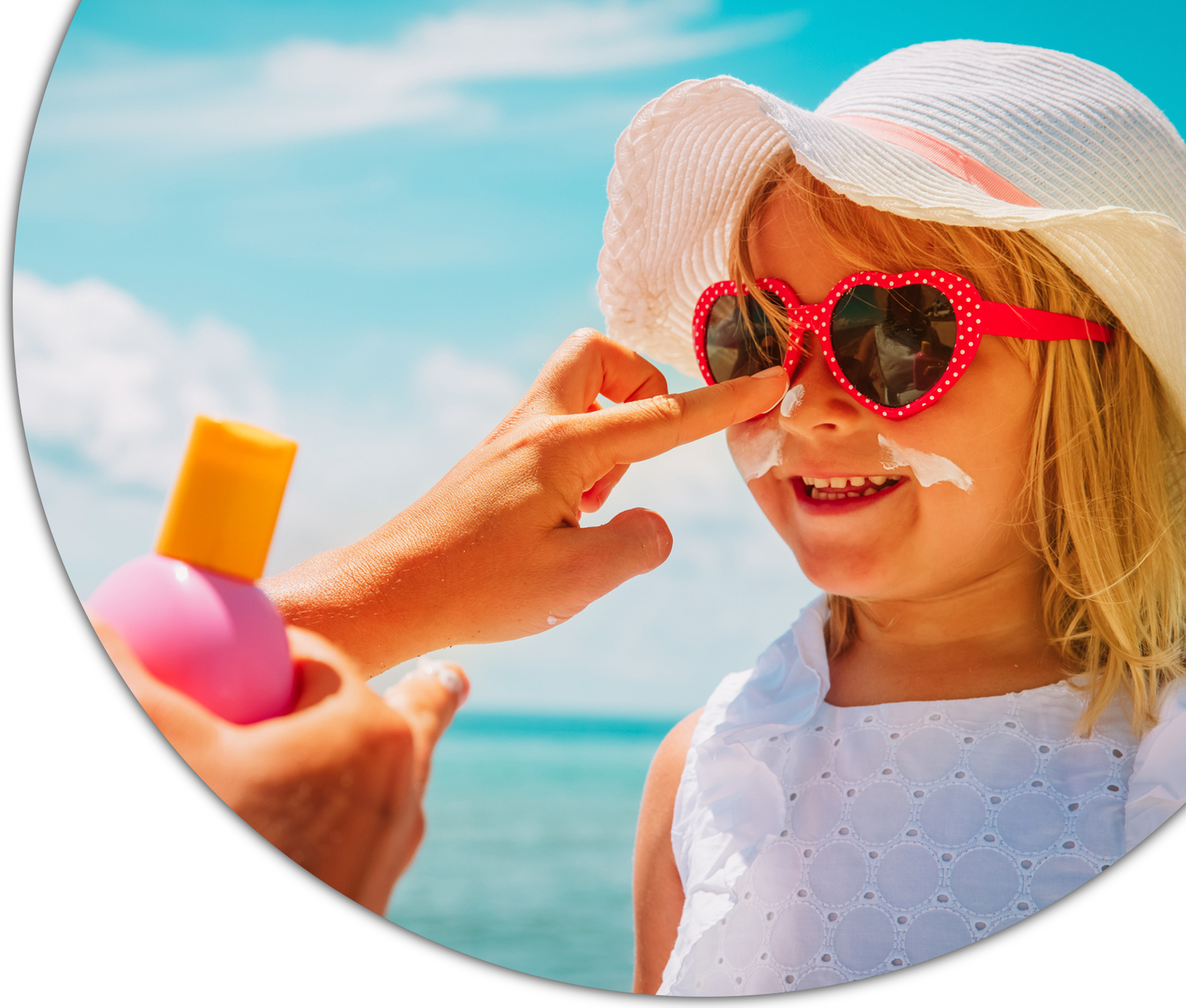 Young girl in heart-shaped sunglasses and a sun hat smiles while sunscreen is applied to her nose, with the sea and sky behind her.